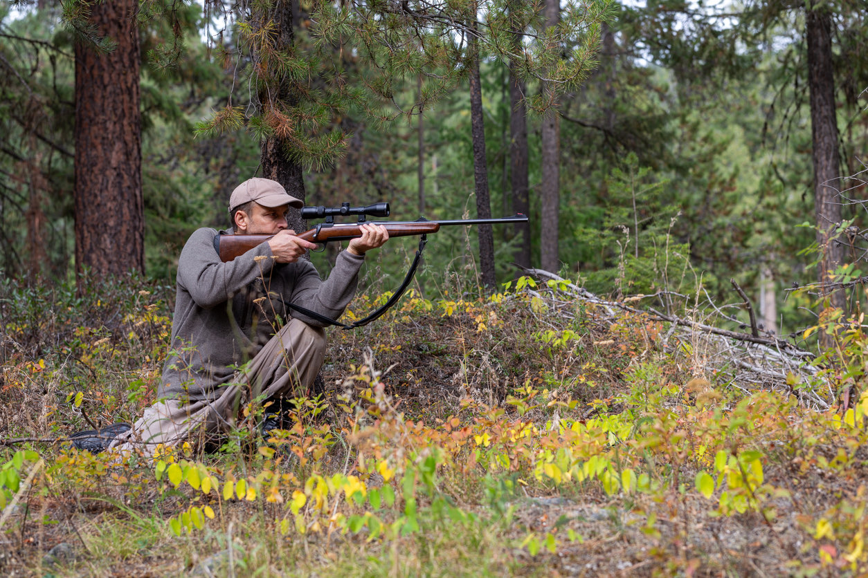 Hunter kneeling in the woods and aiming a scoped rifle
